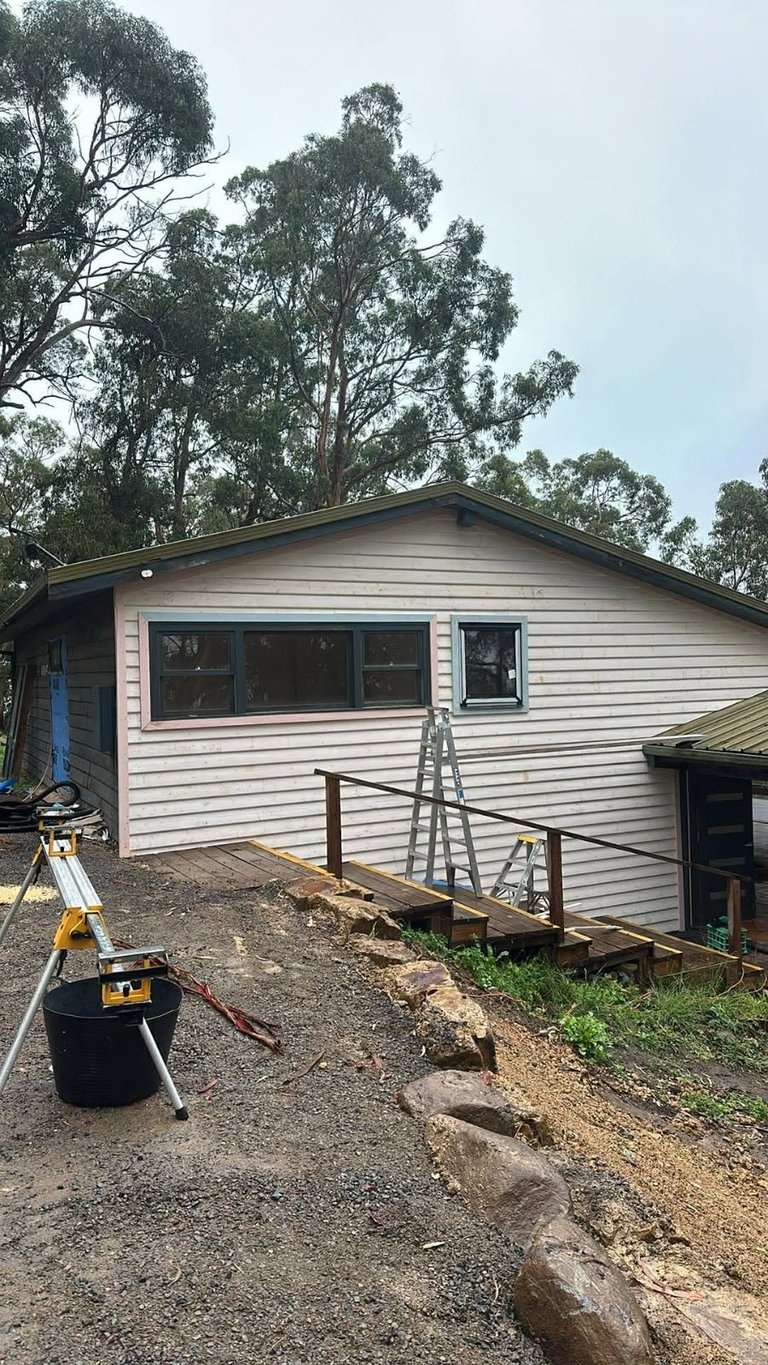 A white residential house with large windows on a hillside, surrounded by tall eucalyptus trees, with construction equipment and materials visible in the yard.